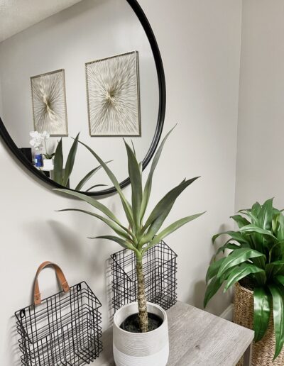 A modern corner with a potted plant, a round mirror, decorative wall art, and a basket with accessories.