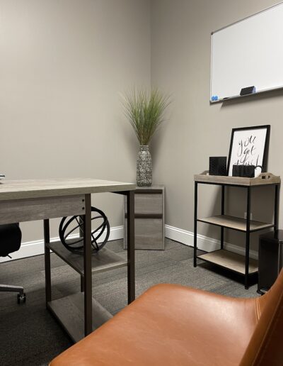 A modern office corner with a desk, brown chair, shelving unit, decorative plant, and a framed motivational quote.