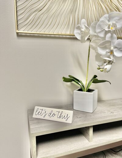 A motivational sign saying "let's do this" placed on a wooden shelf beside a potted orchid, with decorative wall art in the background.