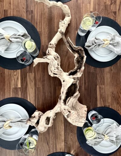A top-down view of a dining table set with black plates, silver napkins, and glasses of water with lemon slices, accented by a natural wood centerpiece.