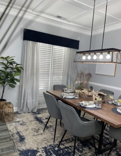A modern dining room featuring a wooden table with place settings, surrounded by gray chairs, with a geometric chandelier above, next to a window draped with curtains and a decorative plant in the corner.