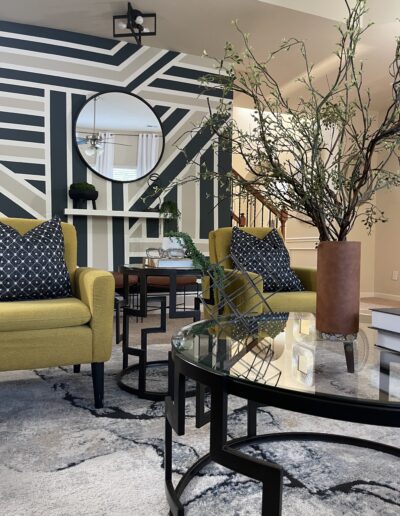 Modern living room interior featuring geometric wallpaper, mustard armchairs, round mirrors, and a glass tabletop with decorative branches.