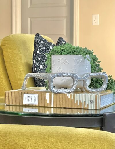 Elegant home decor arrangement featuring a stack of books, a decorative planter with greenery, and eyeglass-shaped ornaments on a glass table with a yellow armchair in the background.