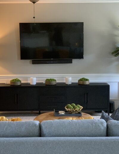 A modern living room with a wall-mounted tv, a black console, decorative plants, and a grey sofa with cushions.