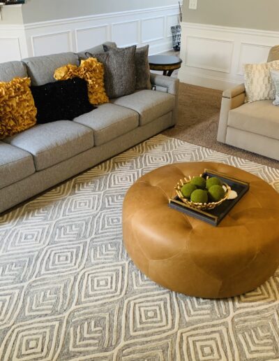 A well-organized living room featuring a gray sectional sofa with yellow accent pillows, a beige armchair, a round leather ottoman, and a geometric patterned rug.