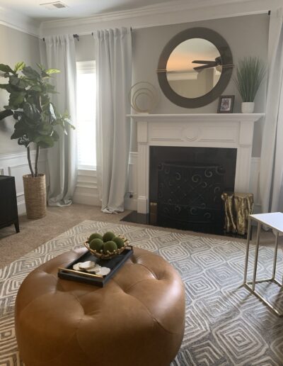 An elegant living room with a neutral color palette, featuring a white fireplace, round mirror, a leather ottoman, and decorative elements.