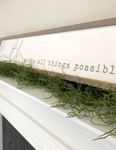 Decorative wooden sign with the quote "faith makes all things possible" on a fireplace mantel, adorned with greenery and a circular metal ornament to the left.