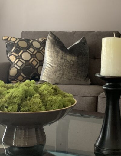 An elegant living room corner with a gray sofa adorned with decorative cushions, a bowl of green moss on a coffee table, and tall candlesticks with white candles.