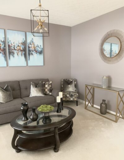 A modern living room featuring a gray sofa with decorative pillows, abstract wall art, a round glass-top coffee table, and a mirrored wall accent.